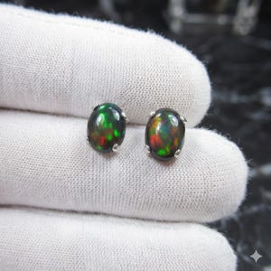 May include: A pair of oval-shaped black opal stud earrings. The opals display a vibrant play-of-color with flashes of green, red, and blue. Each earring is set in a silver-colored metal setting, showcasing the opals' iridescent qualities.
