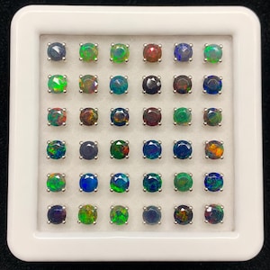 Könnte beinhalten: Ein weißes, quadratisches Display-Etui mit sechsunddreißig runden, facettierten Opal-Edelsteinen, die in einem sechs-mal-sechs-Raster angeordnet sind. Jeder einzelne Stein ist in einer silberfarbenen Metallfassung mit Krappen gefasst und zeigt eine Vielzahl schillernder Farben, darunter Blau, Grün, Rot und dunkle Töne.