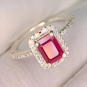 May include: A silver ring featuring a rectangular, deep red center stone set in a halo of small, clear, sparkling stones. The band is thin and also encrusted with a row of tiny, clear, brilliant-cut stones along the visible upper shank.