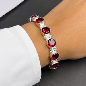 Könnte beinhalten: Ein silbernes Armband mit ovalen, tiefroten Edelsteinen. Die Edelsteine sind in einzelnen Silberfassungen gefasst und durch Silberglieder verbunden. Das Armband wird an einem Handgelenk gezeigt, um das Design und die Farbe des Schmucks hervorzuheben.