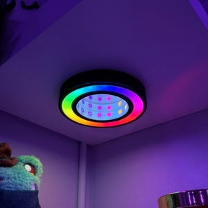 May include: A black, circular LED light fitting with a rainbow-coloured outer ring and a series of small, coloured lights in the centre. The light is mounted on a white shelf, casting colourful light on the surrounding area. A modern lighting solution.
