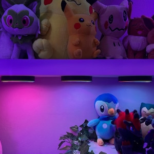 Może przedstawiać: Kolekcja pluszowych zabawek, w tym Pikachu, Sprigatito, Mimikyu, Eevee, Piplup, Snorlax i inne, ułożone na dw&oacute;ch p&oacute;łkach. Trzy okrągłe czarne lampki są zamontowane pod g&oacute;rną p&oacute;łką, rzucając żywą fioletową i niebieską poświatę na otoczenie.