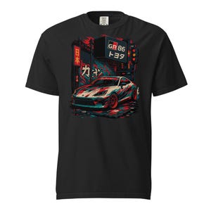 以下が含まれることがあります： 黒いTシャツ。白、赤、青のスポーツカーのグラフィック。日本語のテキストと「GR 86 トヨタ」のサイン。車は赤と青の色合いの都市シーンの背景に設定されています。