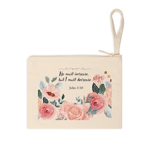 May include: A beige canvas zippered pouch featuring a floral design with pink and peach roses, green leaves, and the text "He must increase, but I must decrease" from John 3:30. The pouch has a beige loop.