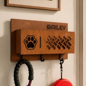May include: A wall-mounted wooden pet leash holder with a rectangular storage bin. The front features a laser-cut paw print, a pattern of small bone shapes, and the name BAILEY engraved in black. Three black metal hooks hang from the bottom.
