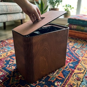 May include: A rectangular waste bin constructed from dark brown wood with a matching lid. The bin features vertical slats along the side edges and a flat top lid with a small handle. A black plastic liner is visible inside the open container.