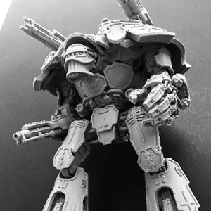 Puede incluir: Miniatura en escala de grises de una máquina de guerra fuertemente blindada, con múltiples cañones y detalles mecánicos intrincados. El modelo tiene una cabeza grande, hombros anchos y piernas poderosas, lo que sugiere un tema de ciencia ficción o fantasía.