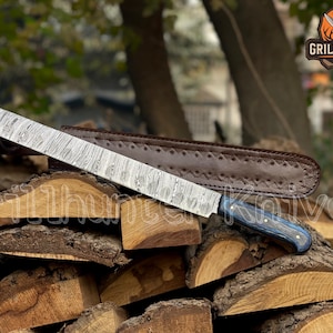 Könnte beinhalten: Ein langes, gemustertes Stahlmesser mit blauem und schwarzem Griff liegt auf einem Holzstapel. Eine braune Lederscheide befindet sich hinter dem Messer. Das Messer hat ein dekoratives Muster auf der Klinge. Das Grill Hunter Knives Logo befindet sich oben rechts.