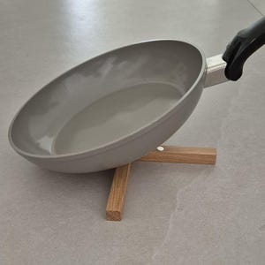 Könnte beinhalten: Eine graue Antihaft-Bratpfanne mit schwarzem Griff, die auf einem kleinen, hellbraunen Holz-Topfuntersetzer ruht. Die Pfanne ist angewinkelt und zeigt ihr Inneres. Der Topfuntersetzer hat ein einfaches, kreuzförmiges Design.