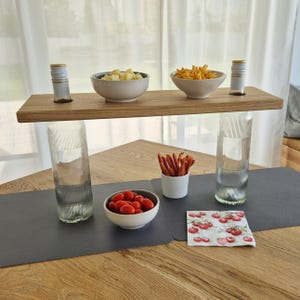 Könnte beinhalten: Ein hölzernes Servierregal mit zwei Flaschen, zwei Schalen mit Snacks und zwei hohen Gläsern. Das Regal wird von den Gläsern getragen. Darunter befinden sich eine Schüssel mit Tomaten, eine kleine Tasse mit Snacks und eine Serviette mit Kirschmuster.