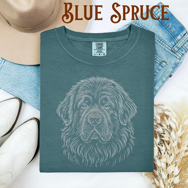May include: Blue Spruce t-shirt with a white line drawing of a Newfoundland dog's face. The shirt has a crew neck and a Comfort Colors tag. The words "Blue Spruce" are written in a decorative font at the top of the image.