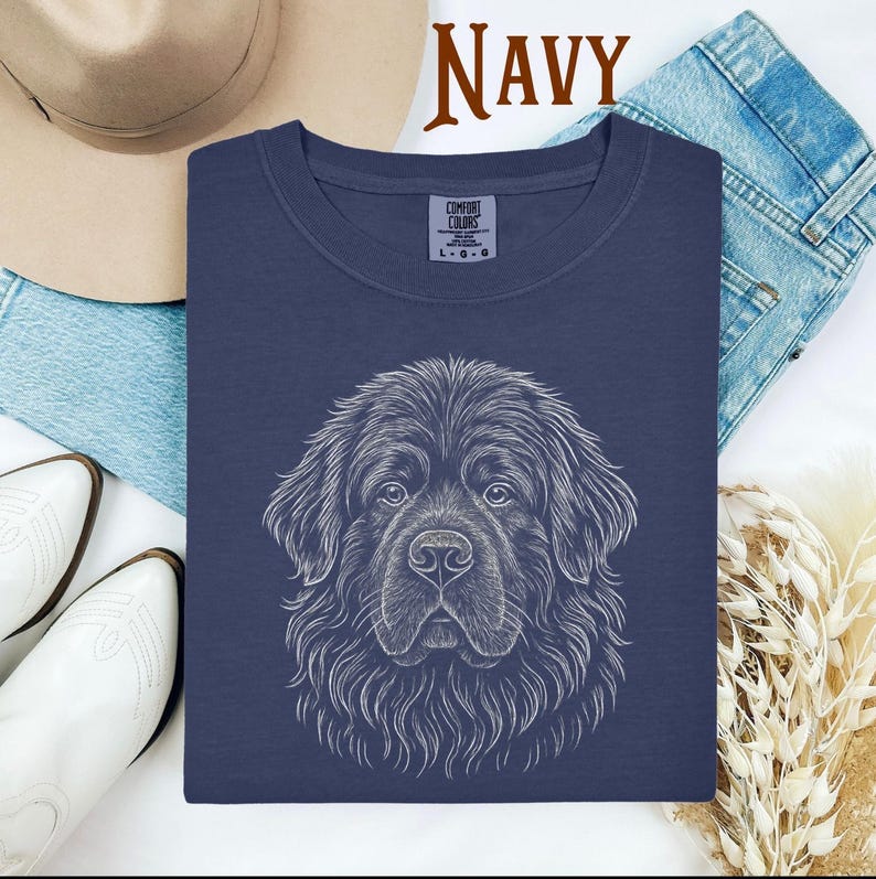 May include: Navy blue t-shirt with a detailed white line drawing of a Newfoundland dog's face. The word "NAVY" is printed in brown at the top. The shirt has a Comfort Colors tag. The shirt is surrounded by a hat, jeans, and boots.