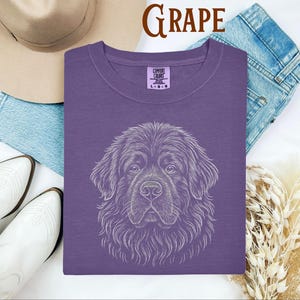 May include: A grape-colored t-shirt with a white line drawing of a dog's face. The word "GRAPE" is printed in brown at the top. The shirt is next to a pair of blue jeans, white boots, and a tan hat.