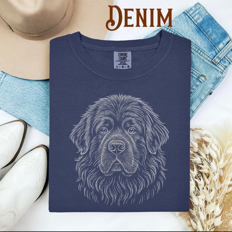May include: Navy blue Comfort Colors sweatshirt featuring a detailed white line drawing of a Newfoundland dog's face. The word "DENIM" is in a brown script font at the top. The sweatshirt is surrounded by a hat, jeans, and white boots.
