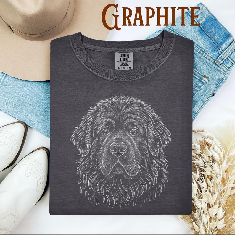 May include: Graphite-colored t-shirt featuring a detailed white line drawing of a Newfoundland dog's face. The shirt has a crew neck and a Comfort Colors tag. The word "Graphite" is printed in brown at the top. The shirt is displayed with jeans, a hat, and boots.