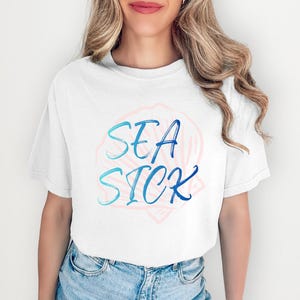 Puede incluir: Camiseta blanca con las palabras "SEA SICK" en escritura azul, sobre un diseño de concha rosa claro. La camiseta es de estilo casual, adecuada para el uso diario.