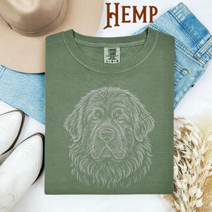 May include: Sage green Comfort Colors t-shirt featuring a detailed white line drawing of a Newfoundland dog's face. The word "HEMP" is printed in brown at the top. The shirt is displayed with a hat, jeans, and boots.