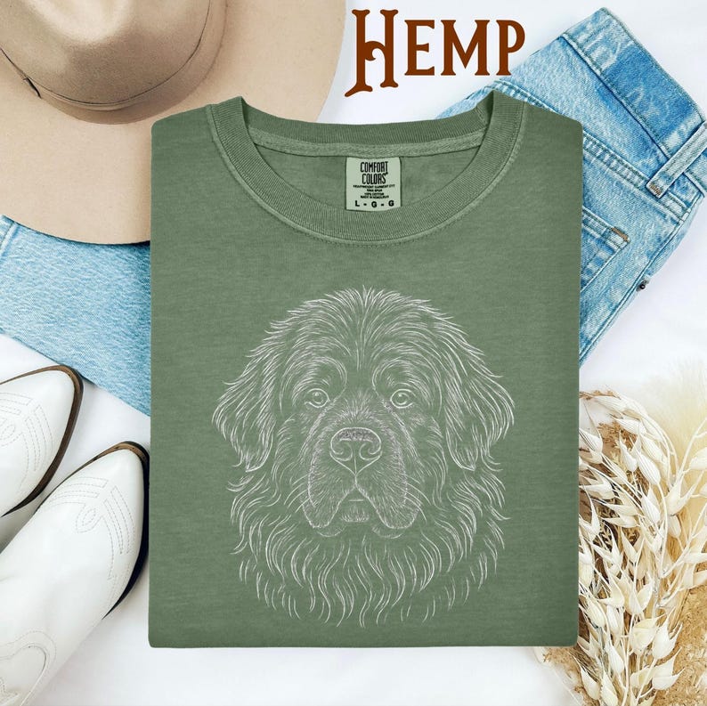 May include: Sage green Comfort Colors t-shirt featuring a detailed white line drawing of a Newfoundland dog's face. The word "HEMP" is printed in brown at the top. The shirt is displayed with a hat, jeans, and boots.