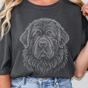 May include: Dark gray t-shirt featuring a detailed, white line drawing of a Newfoundland dog's face. The dog's fur is depicted with intricate lines, creating a textured effect. The shirt has a classic crew neck and short sleeves.