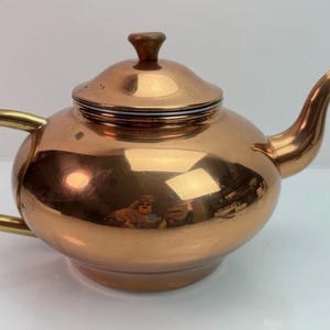 May include: A polished copper teapot featuring a rounded body, a curved spout, and a brass handle. The teapot includes a matching lid with a small knob on top. The metallic surface reflects the surrounding environment, showing a warm, reflective finish.