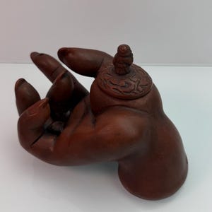 May include: A dark brown carved wooden sculpture shaped like an open hand with fingers curled upward. A circular lid with intricate swirling patterns and a small figure on top rests on the palm area, creating a container with a removable cover.