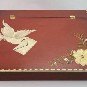 May include: A rectangular wooden writing desk box with a dark red finish. The lid features a hand-painted white dove carrying an envelope on the left and a yellow flower with green leaves on the right. The edges have a scalloped gold trim.