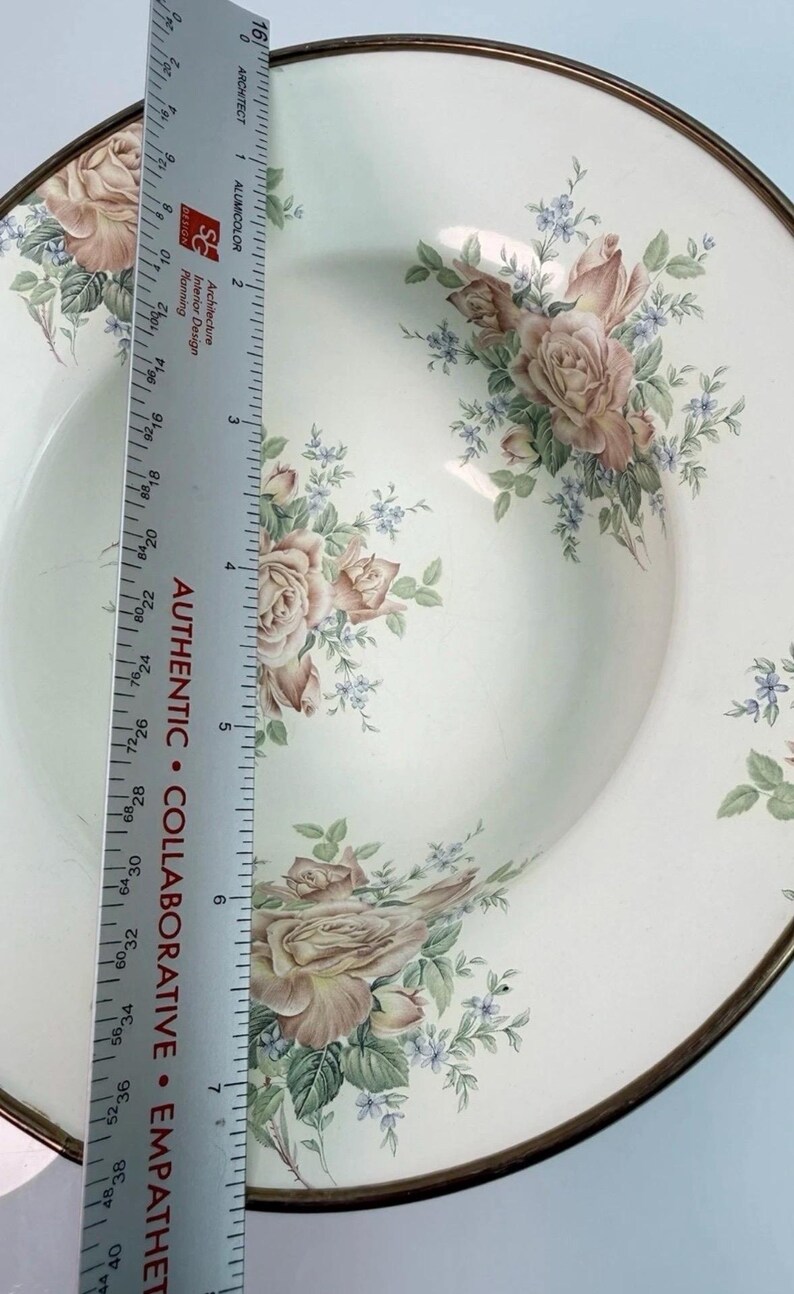 K&ouml;nnte beinhalten: Eine runde Keramikschale mit cremefarbenem Grund und einem Blumenmuster aus rosa Rosen und kleinen blauen Bl&uuml;ten. Ein Metalllineal liegt auf der Schale und zeigt einen Durchmesser von etwa 19 cm mit einem metallischen Rand.