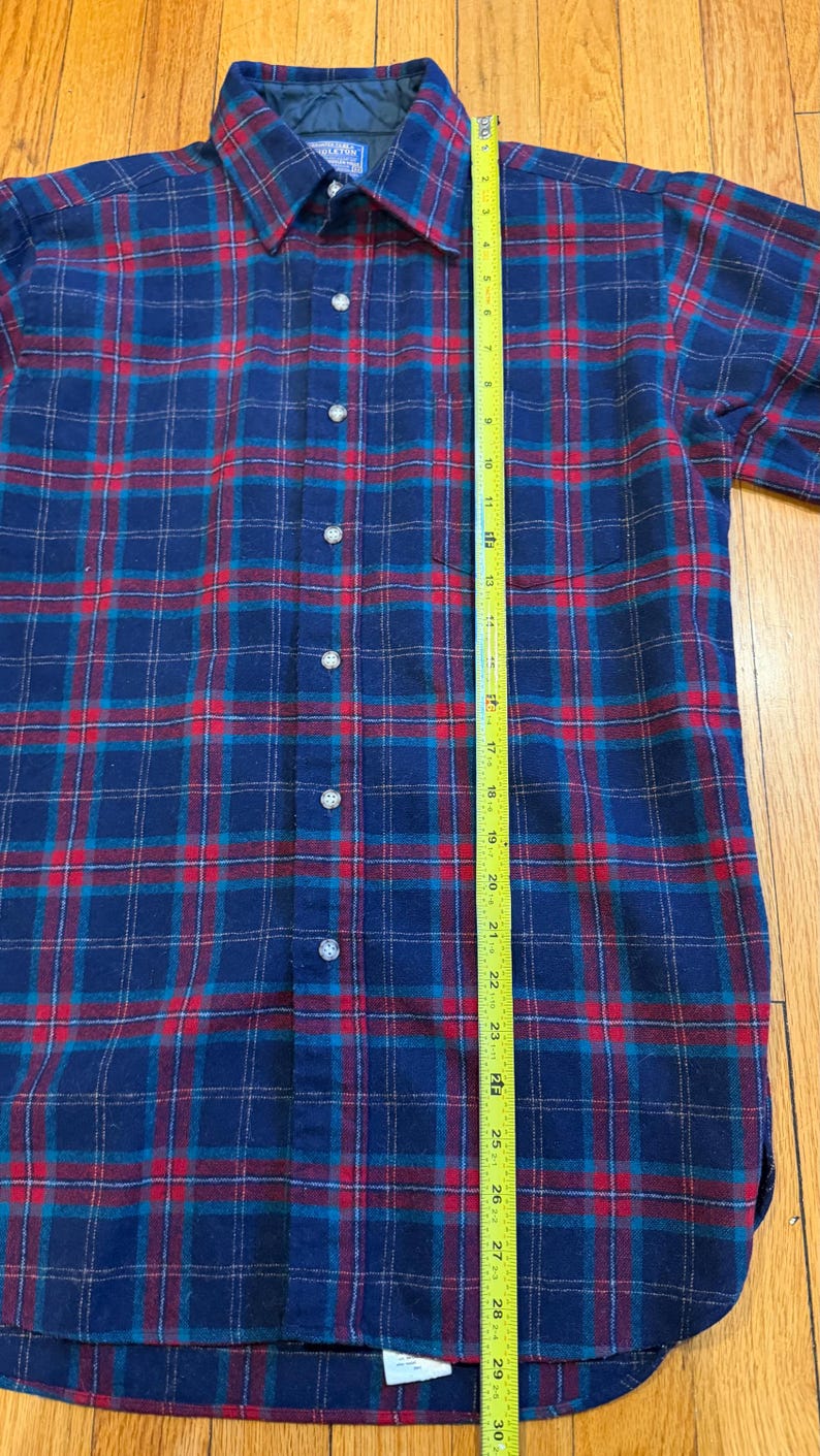 Puede incluir: Una camisa abotonada a cuadros azules, rojos y turquesas. La camisa mide aproximadamente 76 cm de largo, medida con una cinta m&eacute;trica amarilla. La camisa tiene cuello y pu&ntilde;os abotonados. La camisa est&aacute; hecha de un material suave, similar a la franela.