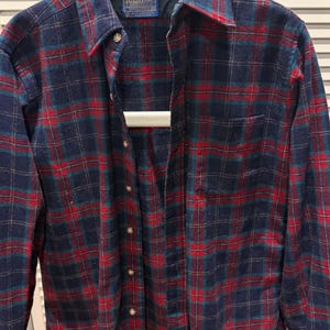 Puede incluir: Una camisa de franela a cuadros azules, rojos y verdes. La camisa tiene una parte delantera con botones, un cuello y un bolsillo en el pecho. La etiqueta Pendleton es visible en la parte superior izquierda. La camisa est&aacute; colgada en una percha blanca.