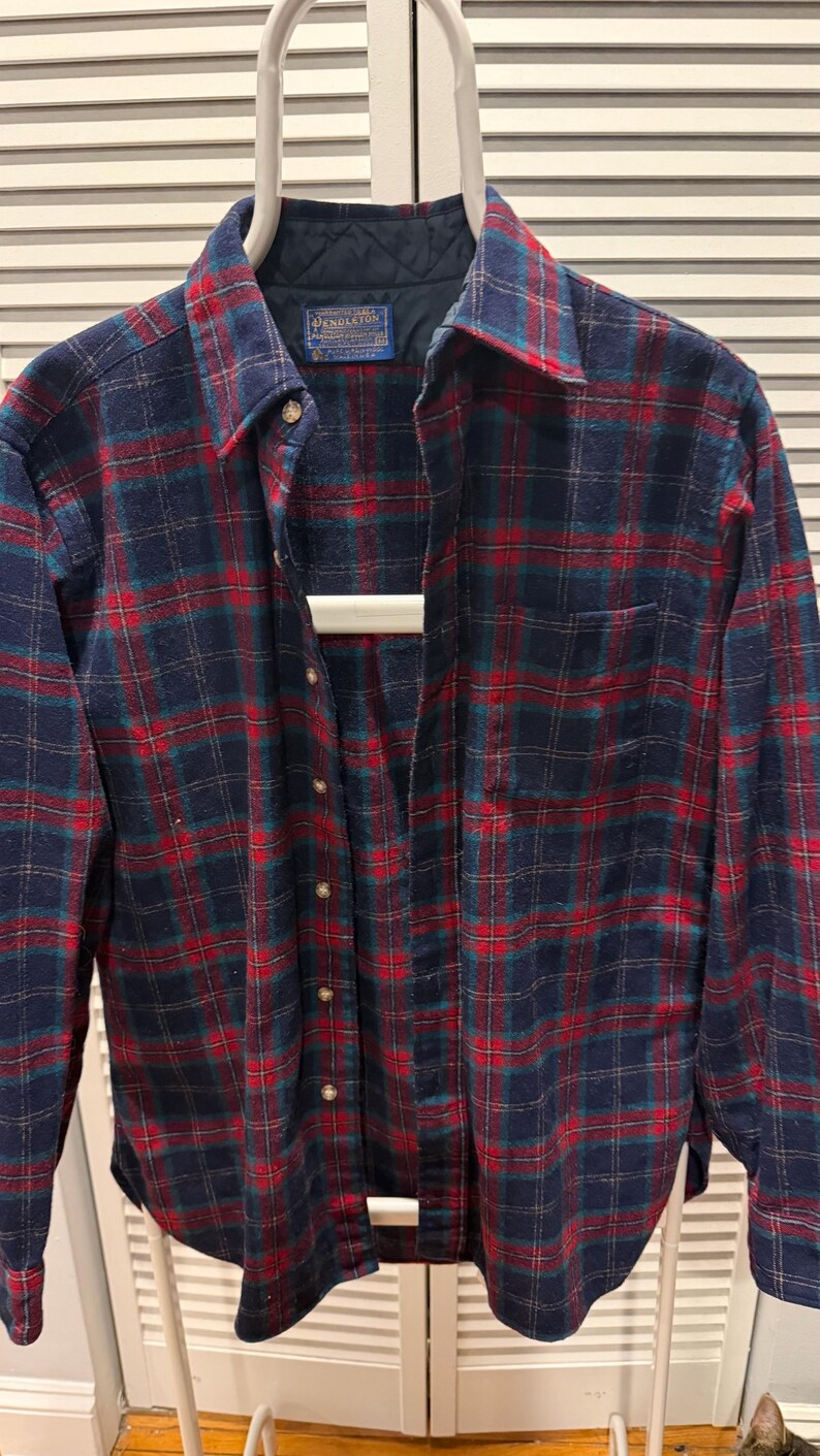 Puede incluir: Una camisa de franela a cuadros azules, rojos y verdes. La camisa tiene una parte delantera con botones, un cuello y un bolsillo en el pecho. La etiqueta Pendleton es visible en la parte superior izquierda. La camisa est&aacute; colgada en una percha blanca.
