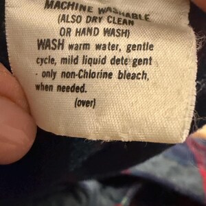 Puede incluir: Primer plano de una etiqueta de tela blanca con texto negro. La etiqueta dice "MACHINE WASHABLE (ALSO DRY CLEAN OR HAND WASH)". Proporciona instrucciones de lavado: agua tibia, ciclo suave, detergente suave y lej&iacute;a sin cloro cuando sea necesario.