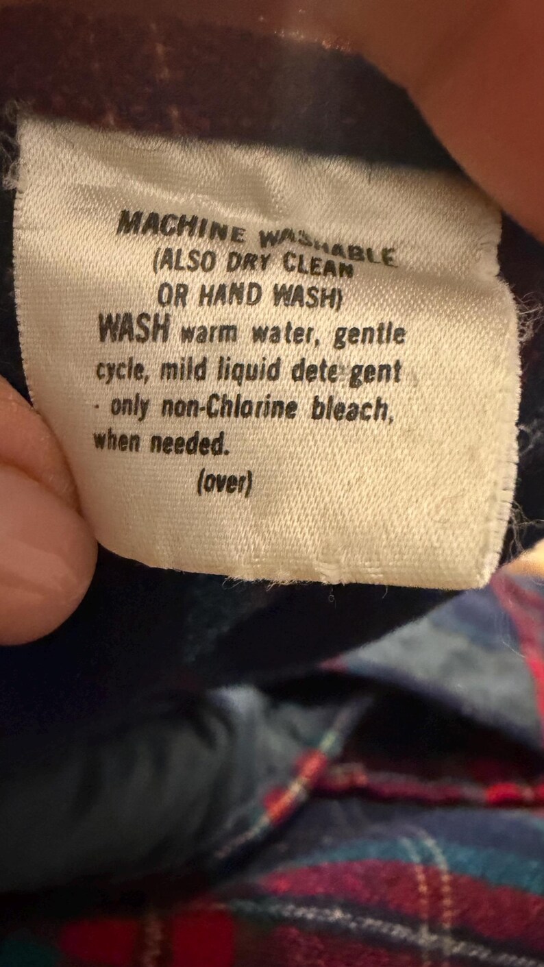 Puede incluir: Primer plano de una etiqueta de tela blanca con texto negro. La etiqueta dice "MACHINE WASHABLE (ALSO DRY CLEAN OR HAND WASH)". Proporciona instrucciones de lavado: agua tibia, ciclo suave, detergente suave y lej&iacute;a sin cloro cuando sea necesario.