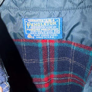 Puede incluir: Primer plano de una chaqueta de lana a cuadros azules y rojos con forro azul acolchado. La chaqueta tiene una etiqueta que dice "Pendleton Woolen Mills, Made in USA". La chaqueta est&aacute; hecha de 100% lana virgen.