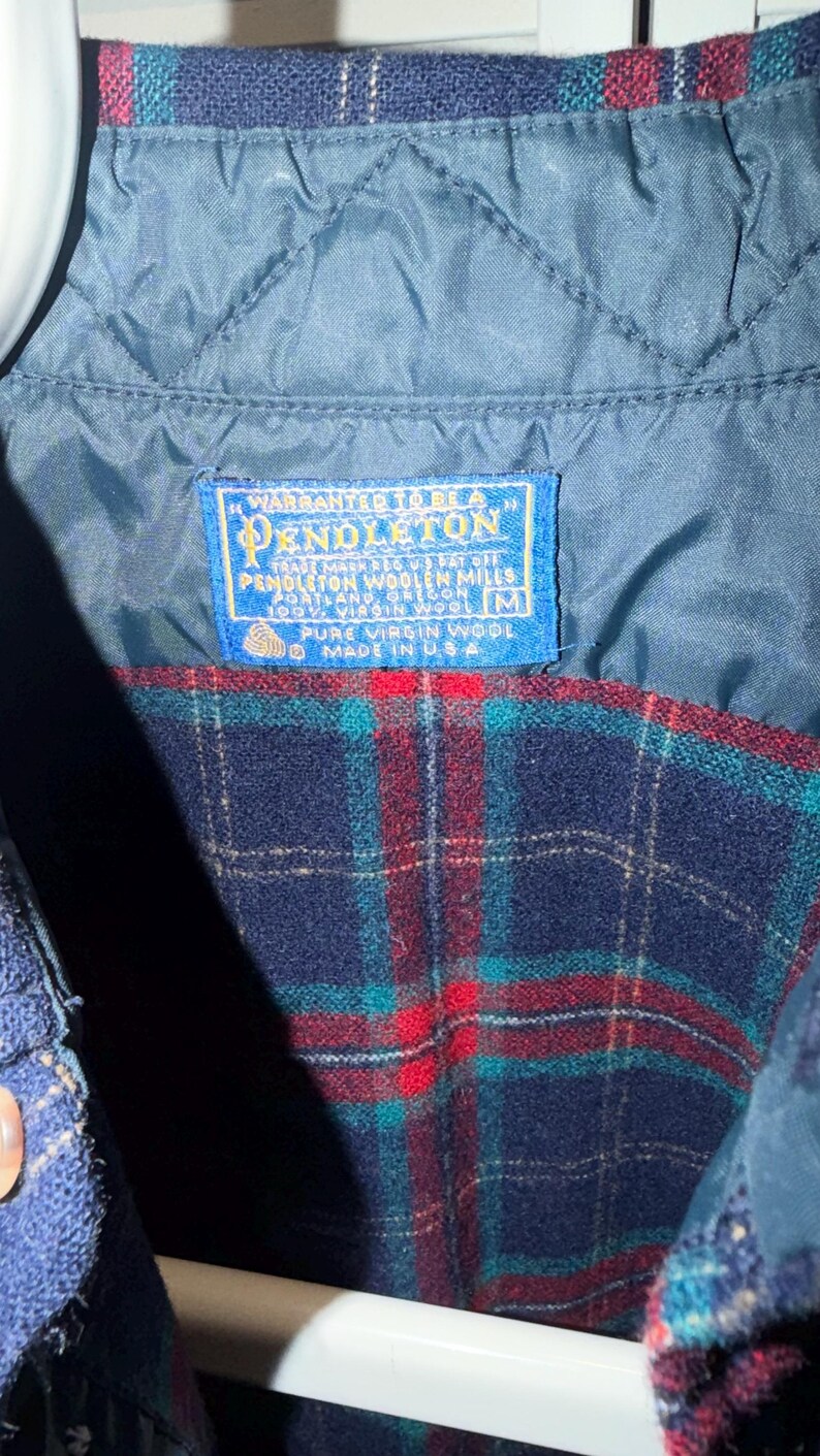 Puede incluir: Primer plano de una chaqueta de lana a cuadros azules y rojos con forro azul acolchado. La chaqueta tiene una etiqueta que dice "Pendleton Woolen Mills, Made in USA". La chaqueta est&aacute; hecha de 100% lana virgen.
