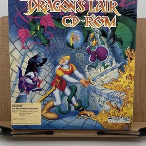 May include: A CD-ROM box for the game Dragon's Lair. The box art features a knight with a sword, dragons, and a princess. The game is for Macintosh computers. The box is blue with colorful illustrations.