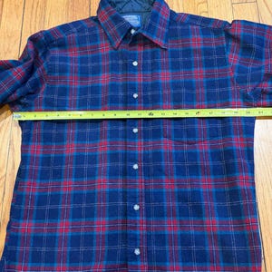 Puede incluir: Camisa de franela a cuadros azules y rojos con botones y mangas largas. La camisa tiene un cuello cl&aacute;sico. Una cinta m&eacute;trica est&aacute; colocada sobre la camisa. La camisa mide aproximadamente 53 cm de ancho.