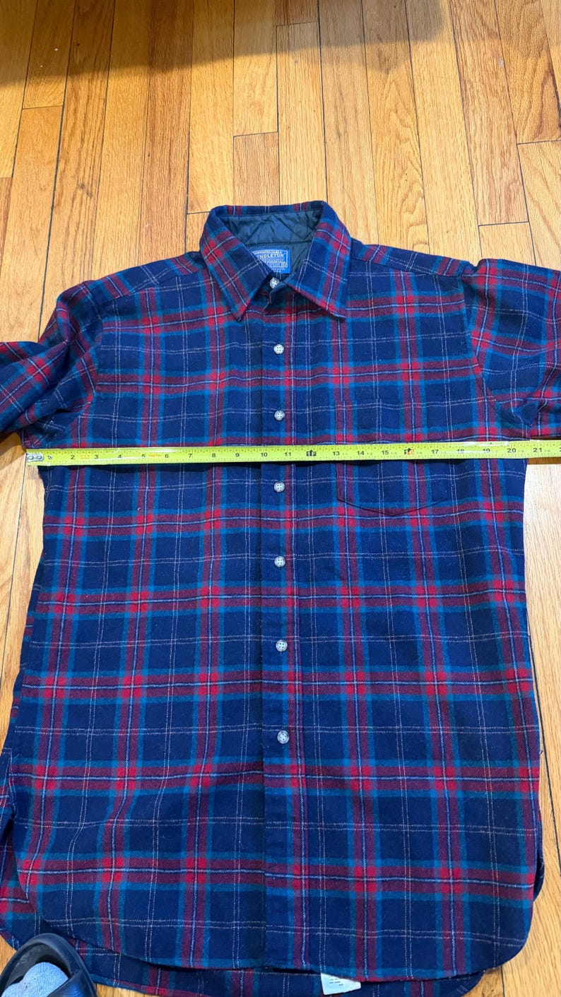 Puede incluir: Camisa de franela a cuadros azules y rojos con botones y mangas largas. La camisa tiene un cuello cl&aacute;sico. Una cinta m&eacute;trica est&aacute; colocada sobre la camisa. La camisa mide aproximadamente 53 cm de ancho.