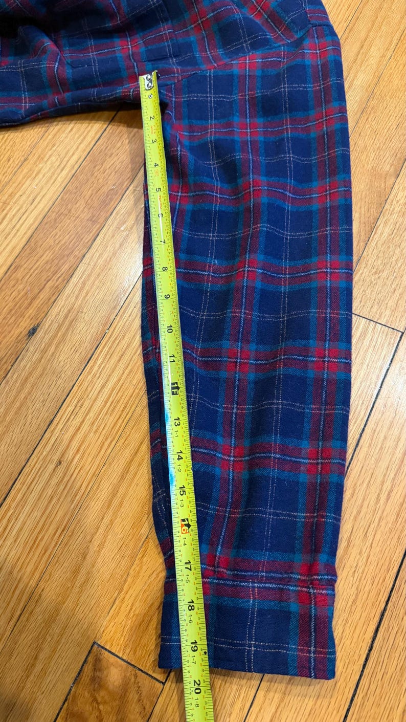 Puede incluir: Una manga de camisa de franela a cuadros azules, rojos y turquesas se mide con una cinta m&eacute;trica amarilla. La manga mide aproximadamente 51 cm de largo. El patr&oacute;n de cuadros es un dise&ntilde;o cl&aacute;sico.