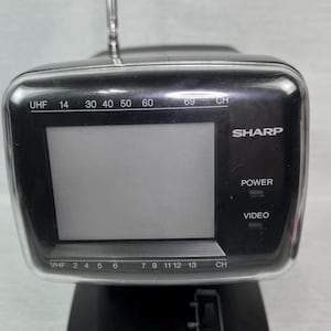 May include: A black Sharp portable TV with a silver antenna. The TV has a screen, channel selection buttons, and power and video buttons. The model has UHF and VHF channel options.