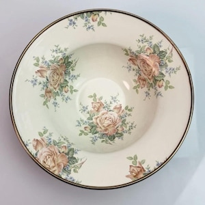 K&ouml;nnte beinhalten: Eine runde Keramikschale mit cremefarbener Basis und einem Blumenmuster aus zartrosa Rosen und kleinen blauen Bl&uuml;ten. Die Schale hat einen breiten Rand und eine d&uuml;nne dunkelbraune oder schwarze Linie entlang der Au&szlig;enkante als Abschluss.