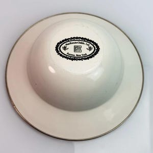 K&ouml;nnte beinhalten: Eine cremewei&szlig;e, abgerundete Sch&uuml;ssel mit einem breiten, flachen Rand und einer metallisch silberfarbenen Kante. Die Unterseite zeigt einen schwarzen ovalen Stempel mit dem Text MacKenzie-Childs, Ltd. und Aurora, New York, umrandet von einem floralen Muster.