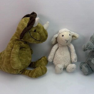 May include: A set of three plush toys including a textured olive green dinosaur with a dark brown crest, a cream-colored lamb with floppy ears, and a small gray elephant with a trunk, all positioned together on a plain white surface.