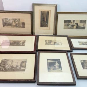 May include: A collection of eight framed prints featuring monochromatic interior scenes and figures, each housed in rectangular frames of varying sizes with wooden or metallic finishes, off-white matting, and glass covers arranged in a group.