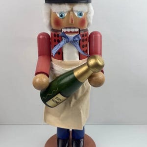 May include: A wooden nutcracker in the form of a chef, wearing a brown beret, red jacket, and apron, holding a green bottle of "DRY WINE". The nutcracker has a blue bow tie and is standing on a wooden base.