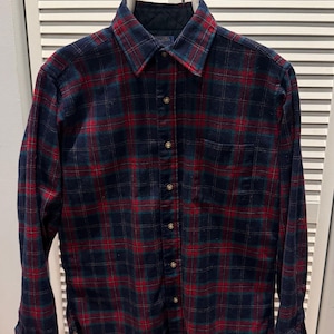 Puede incluir: Una camisa de franela abotonada de manga larga con un patr&oacute;n de cuadros cl&aacute;sico. La camisa presenta una base azul marino con l&iacute;neas de cuadros rojas y verdes. Cuelga de una percha blanca contra una puerta de listones blancos.