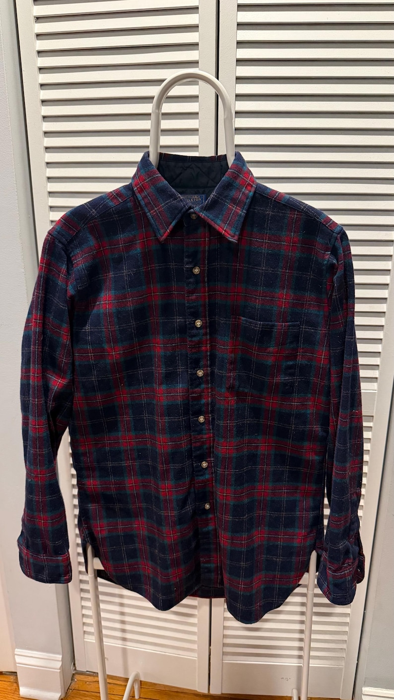 Puede incluir: Una camisa de franela abotonada de manga larga con un patr&oacute;n de cuadros cl&aacute;sico. La camisa presenta una base azul marino con l&iacute;neas de cuadros rojas y verdes. Cuelga de una percha blanca contra una puerta de listones blancos.