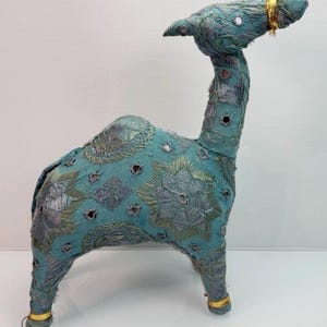 May include: A teal fabric camel figurine featuring intricate silver and gold thread embroidery in geometric star patterns. The surface is embellished with small circular mirrors and gold bands around the neck and legs.