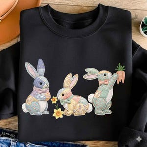 Op de afbeelding: Zwart sweatshirt met drie patchwork konijnen met bloemen- en stippenpatronen. Eén konijn houdt een wortel vast, een ander een bloem en de derde een ei. De konijnen dragen strikjes en staan tegen een zwarte achtergrond.
