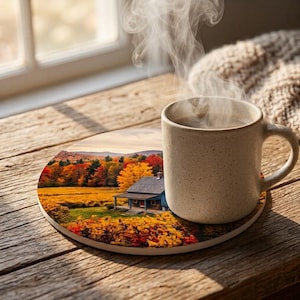 Könnte beinhalten: Ein runder Untersetzer mit einem aufgedruckten Bild eines blauen Hauses, umgeben von herbstlichem Laub in Gelb-, Orange- und Rottönen. Eine beige Keramiktasse mit sichtbarem Dampf, der aus der Öffnung aufsteigt, steht auf der rechten Seite.