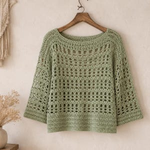 May include: A pale green, open-weave crochet top with a round neckline and three-quarter sleeves. The top is suspended on a wooden hanger. The crochet pattern has a grid-like design. The background is neutral.